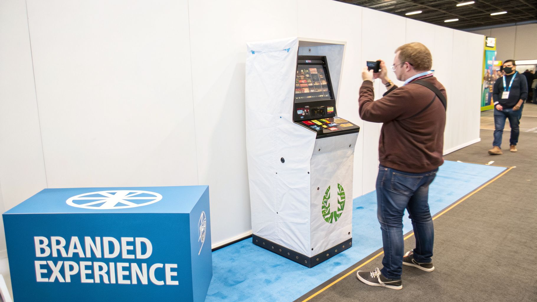 A man photographs a white arcade game machine at an event booth next to a blue branded sign.