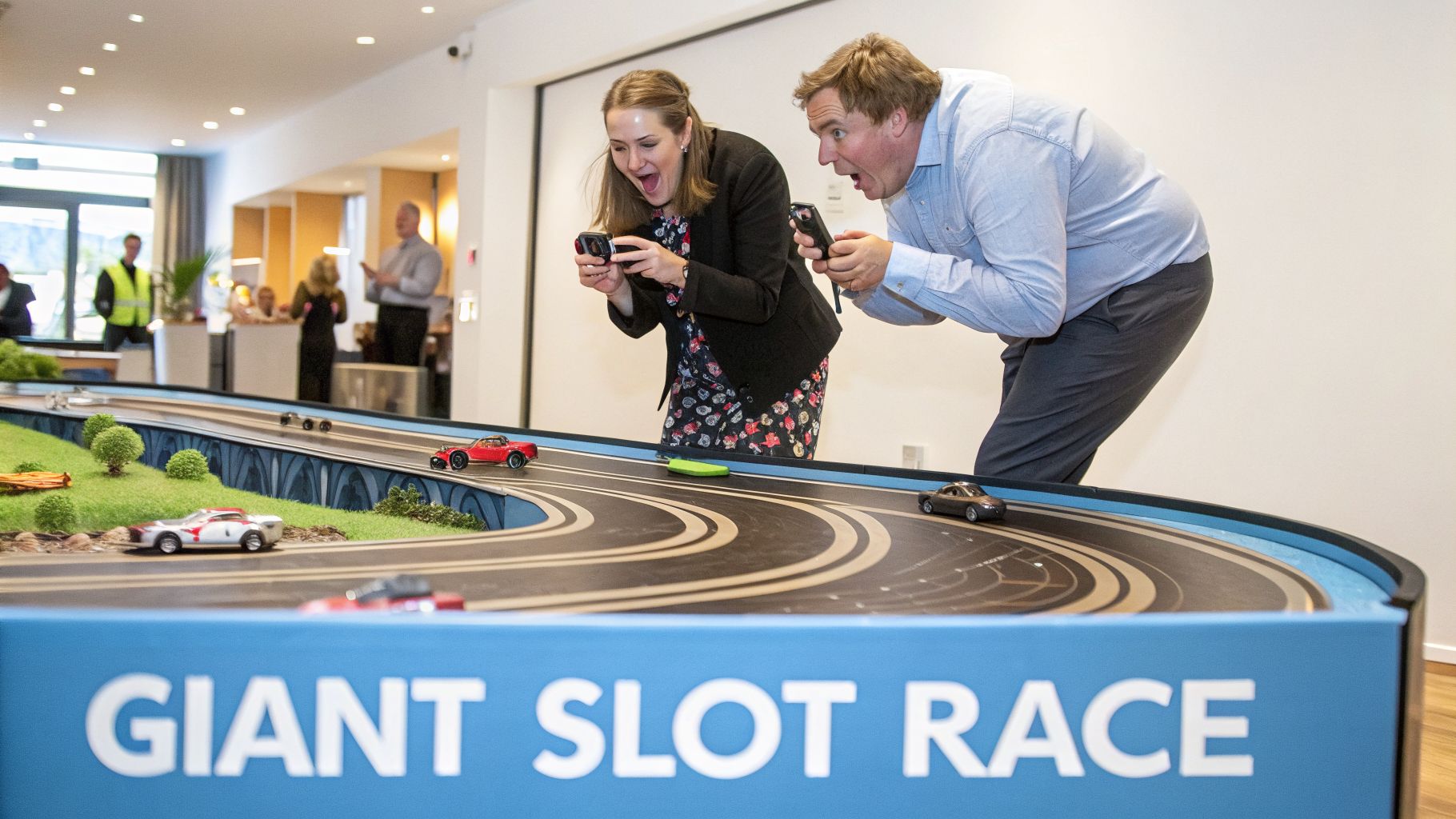 Two adults excitedly play a giant slot car race, controllers in hand, with toy cars on the track.