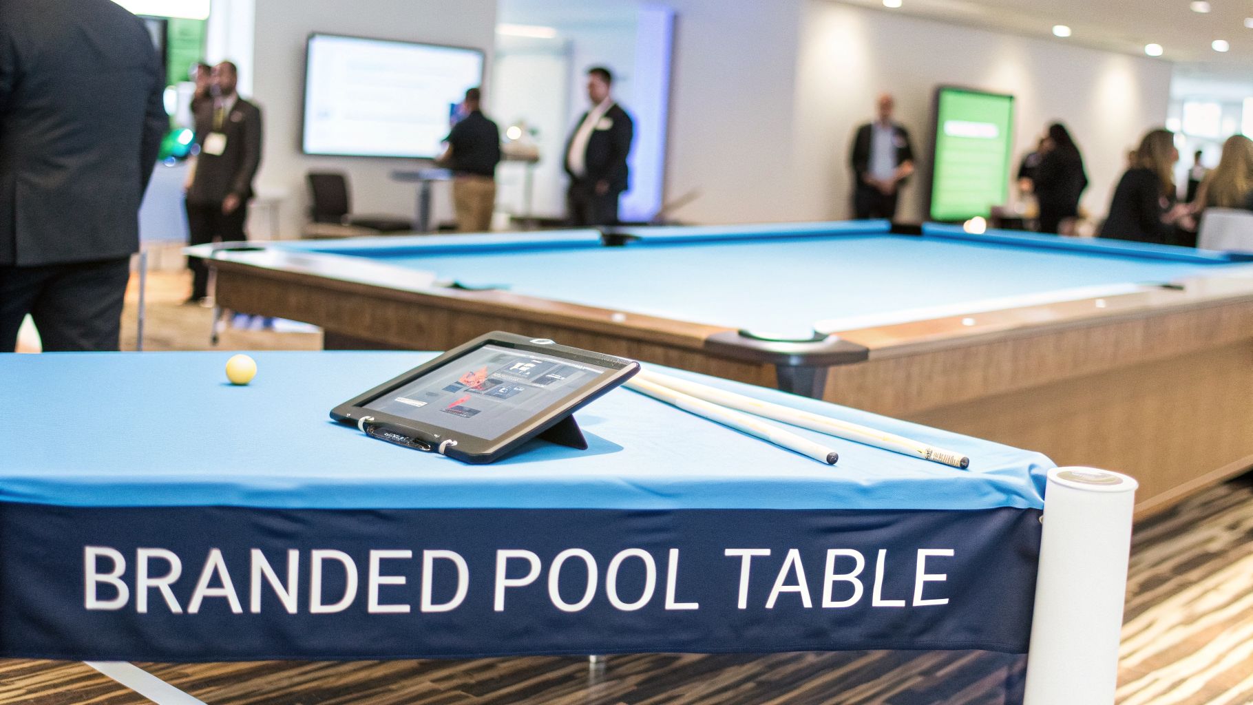 Close-up of a branded pool table with an iPad and cues, people in the background.