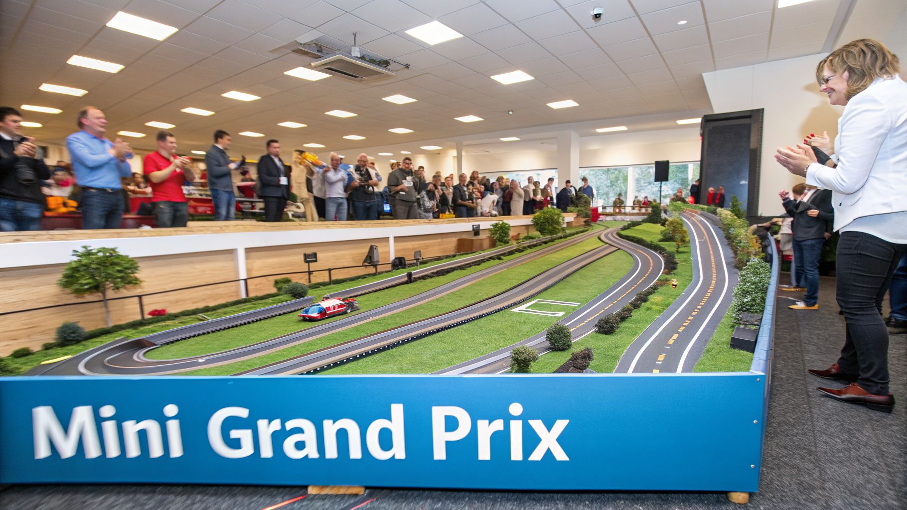 People watch and clap at a large, detailed slot car track with miniature cars and trees.