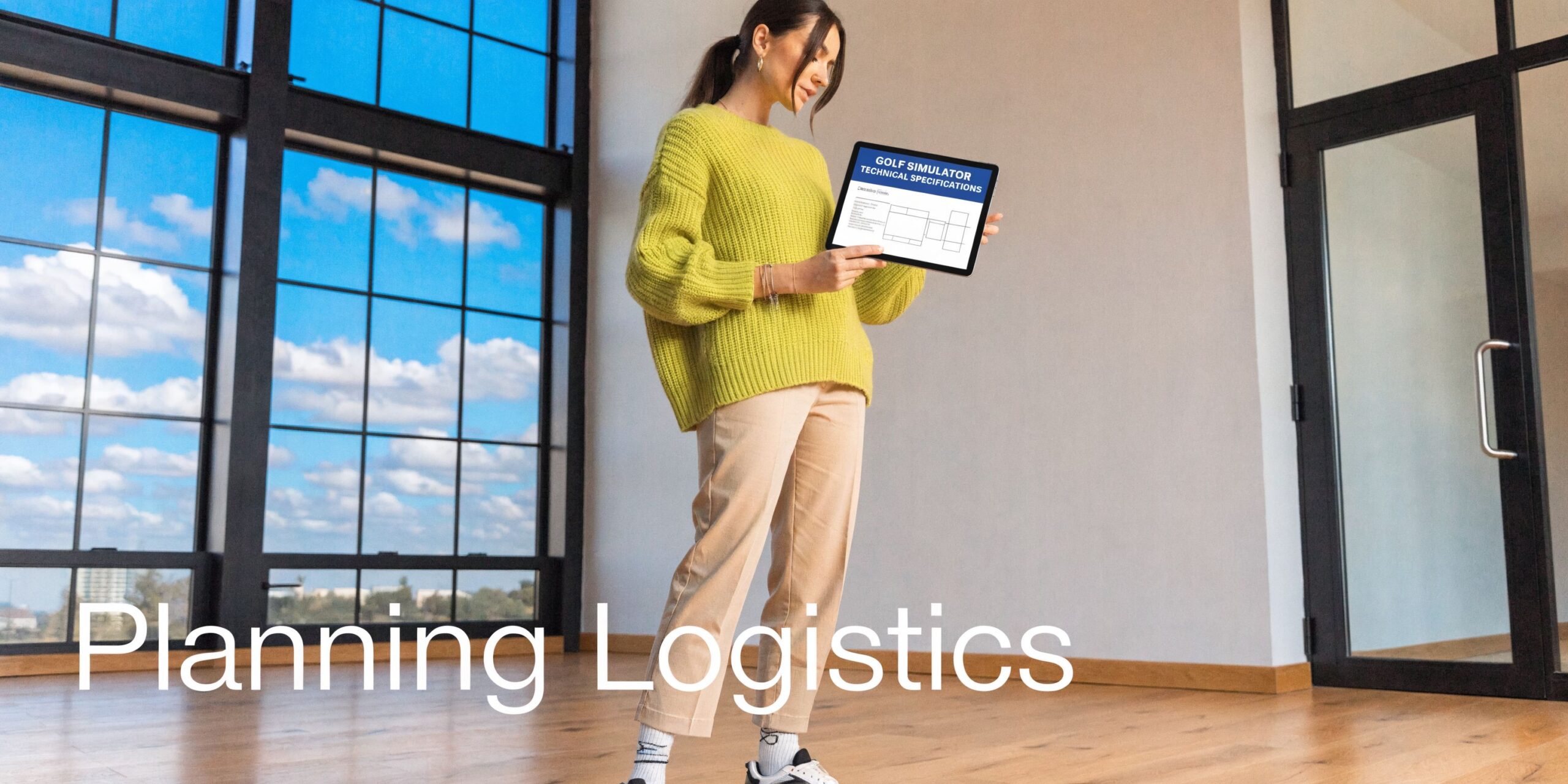 A professional woman standing in an empty room holding a tablet displaying golf simulator technical specifications.