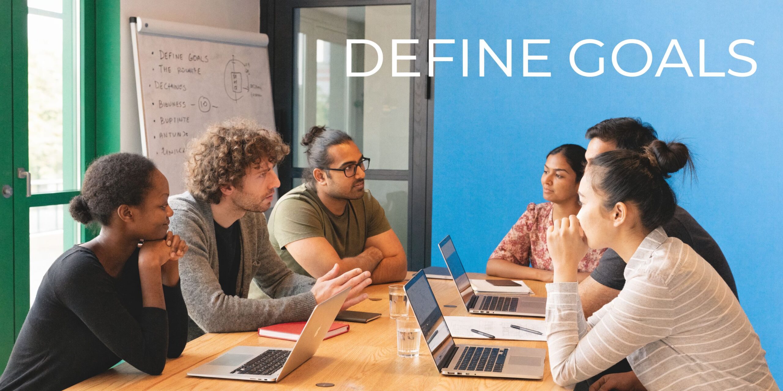 A diverse group of colleagues sitting around a wooden table in an office discussing business goals together.