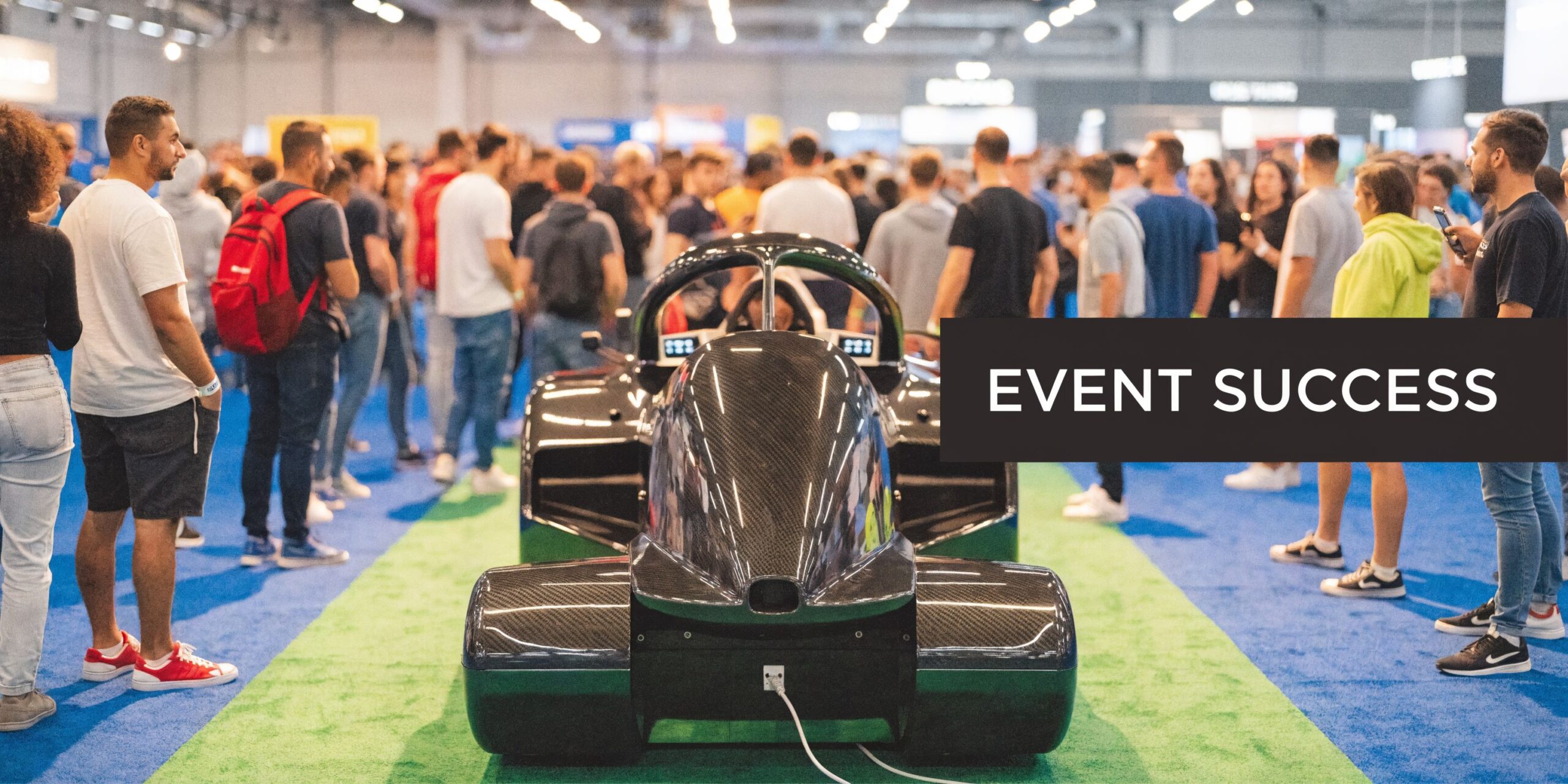 Attendees surrounding a sleek carbon fiber racing simulator at a busy exhibition event on a convention floor.