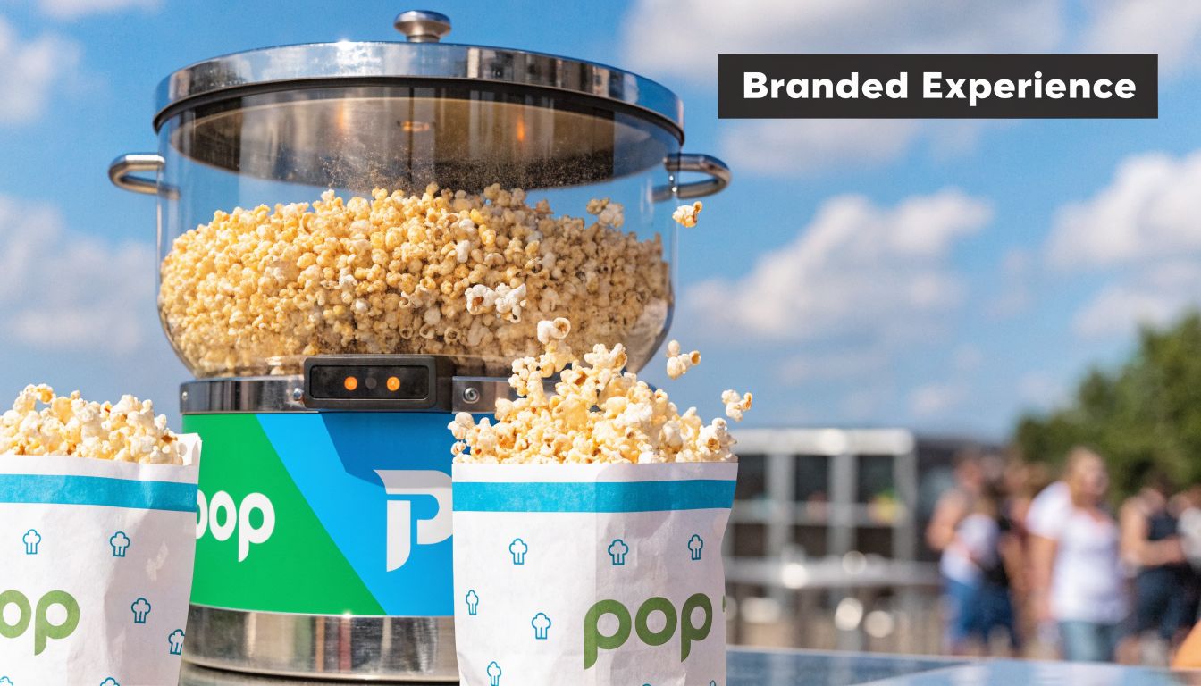 A popcorn machine branded with a logo sits outdoors, filling two white paper bags with fresh popcorn.